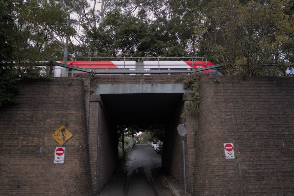 The image shows a narrow underpass beneath a railway bridge constructed of dark brown bricks with a weathered appearance. The underpass is relatively low, with a dark, shadowed interior that obscures the view of what lies beyond. Directly above, a red and white bus is partially visible, parked on a raised roadway with metal railings along its edge. The surrounding environment includes trees with leafy branches extending over the top of the bridge, indicating a semi-urban setting. On either side of the entrance to the underpass, there are two road signs mounted on the brick wall: one yellow diamond-shaped sign indicating a left clearance of 4.2 meters, and two red and white circular no entry signs for vehicles. The surface of the road beneath the bridge appears to be asphalt, with visible tire marks and slight dampness, possibly from recent rain. The scene is lit by natural daylight, creating a neutral tone that highlights the textures of the bricks, metal railings, and foliage, aligning with the context of private or alternative waste handling where clearance restrictions or access limitations might be relevant, such as for rubbish removal or site clearance purposes.