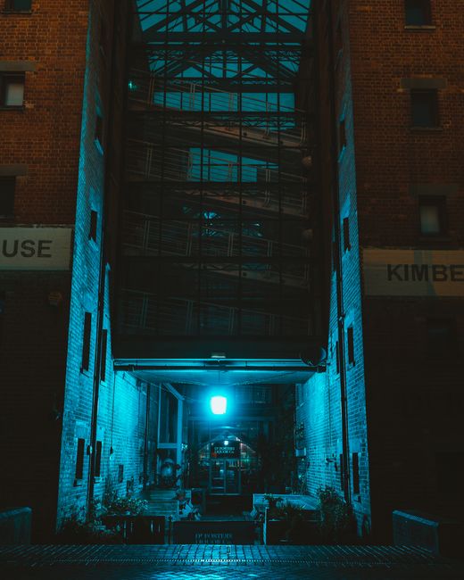 The image depicts a narrow, enclosed urban passageway between two brick buildings, with a metal fire escape staircase visible in the background. The buildings feature a reddish-brown brick finish, and the passage is illuminated by a striking teal-blue light that casts a glow across the brick walls and surrounding area. The ground surface appears to be paved, with some small potted plants positioned along the sides, suggesting a maintained entrance or courtyard area. The fire escape's metallic structure spans the vertical space, with horizontal platforms and ladders visible through the perforated metal. On either side of the entrance to the passage, there are signs attached to the brick facades, partially visible, indicating commercial or building names. The scene evokes urban infrastructure and aesthetic considerations relevant to property access and maintenance, which can be associated with services like private rubbish clearance or on-site waste management, particularly when considering building access in dense city environments. This visual detail aligns with discussions about alternative waste handling methods outside traditional municipal collection routes and highlights the importance of professional clearance. The overall atmosphere is quiet, with subdued lighting emphasizing the structural features and urban setting, appropriate for a professional rubbish removal context involving difficult access points.