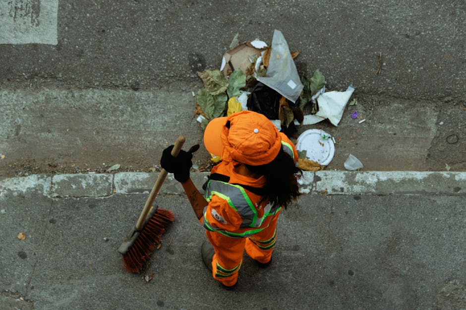 A street cleaner dressed in bright orange high-visibility workwear, including a matching hat and safety vest with reflective stripes, is standing on a paved sidewalk next to a curb. The worker is holding a long-handled broom with a wooden handle and wide red bristles, and is sweeping loose debris and litter from the pathway into a pile near the edge of the pavement. The pile of rubbish consists of various waste items such as crumpled paper, plastic packaging, and a discarded paper plate, some partially folded or crumpled, with textures ranging from smooth plastic to torn paper. The waste is stacked against a plain, slightly weathered concrete wall, which forms the background. The environment appears to be an urban setting, with the street surface showing signs of wear and small scattered debris. This scene illustrates the process of street cleaning or removal of waste which may relate to private waste collection services, as performed by local contractors like rubbishremovalsouthkensington.com, highlighting the importance of maintaining cleanliness and managing rubbish outside residences or commercial premises.