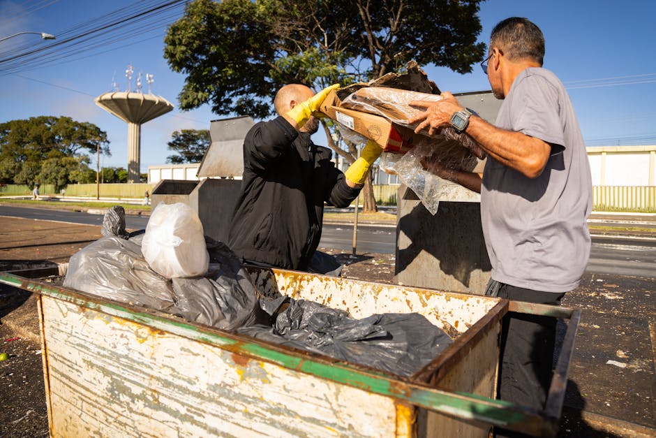 Two men are engaged in a waste collection task outdoors on a bright, clear day. One individual, dressed in a black jacket and yellow gloves, is seen placing a large, dark-colored bag filled with rubbish into an open-top, rusted metal dumpster, which contains several black and grey plastic garbage bags and loose debris. The other man, wearing a light grey t-shirt and glasses, is holding a cardboard box with transparent plastic wrapping, appearing to transfer or inspect items, while standing beside the dumpster. The scene is set on a paved area adjacent to a street with a yellow fence, large green trees, and a water tower visible in the background, indicating a semi-urban environment. Overhead, a clear blue sky and some power lines contribute to the daytime atmosphere. The setup and equipment suggest a service related to private waste removal or on-site clearance, reflecting practices typical of independent rubbish collection services, as seen on www.rubbishremovalsouthkensington.com.