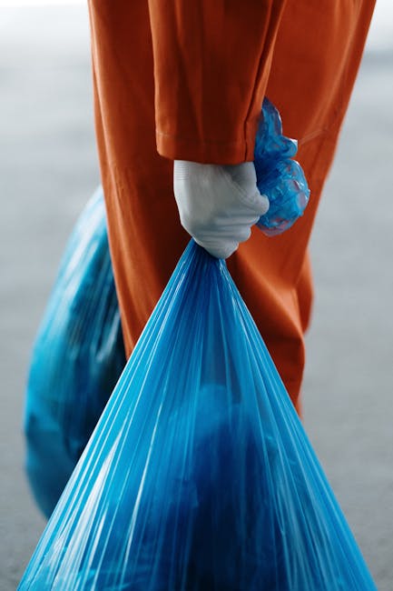 A street cleaner dressed in bright orange high-visibility workwear, including a matching hat and safety vest with reflective stripes, is standing on a paved sidewalk next to a curb. The worker is holding a long-handled broom with a wooden handle and wide red bristles, and is sweeping loose debris and litter from the pathway into a pile near the edge of the pavement. The pile of rubbish consists of various waste items such as crumpled paper, plastic packaging, and a discarded paper plate, some partially folded or crumpled, with textures ranging from smooth plastic to torn paper. The waste is stacked against a plain, slightly weathered concrete wall, which forms the background. The environment appears to be an urban setting, with the street surface showing signs of wear and small scattered debris. This scene illustrates the process of street cleaning or removal of waste which may relate to private waste collection services, as performed by local contractors like rubbishremovalsouthkensington.com, highlighting the importance of maintaining cleanliness and managing rubbish outside residences or commercial premises.