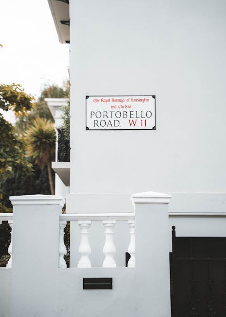A close-up view of the exterior corner of a modern residential building featuring a white wall and a small balcony with decorative balustrades. A rectangular street sign mounted on the wall displays the name 'Portobello Road' and indicates it is under the jurisdiction of the Royal Borough of Kensington and Chelsea, with the postcode prefix W.11. The building's surface has a smooth, painted finish, contrasting with the textured black mailbox inset into the lower part of the wall. In the background, blurred greenery and trees are visible, suggesting the property is situated in a suburban area. Natural daylight illuminates the scene, emphasizing the clean lines and fresh appearance of the structure. The image conveys a typical urban or suburban setting where private properties might handle their rubbish collection independently, aligning with alternative waste disposal options outside local council services. The overall composition showcases the neat, well-maintained exterior indicative of a typical residential environment associated with waste management services like those offered by rubbishremovalsouthkensington.com.