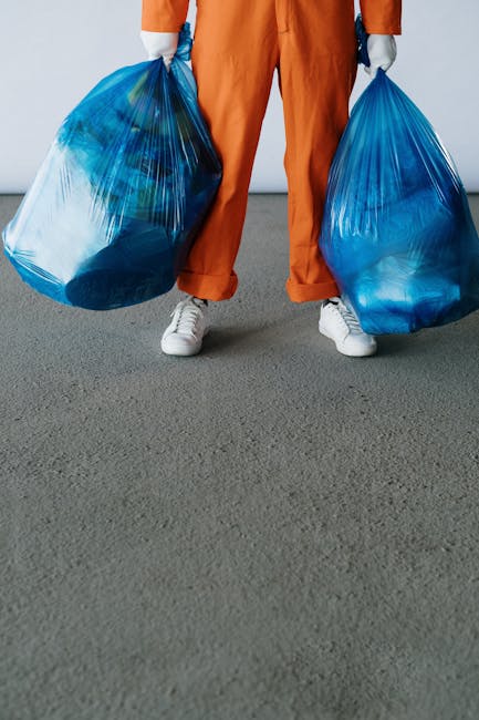 The image shows a person standing on a grey carpeted surface, holding two large blue plastic rubbish bags filled with waste materials. The individual is wearing orange work trousers and white sneakers. The bags are semi-transparent, revealing a mixture of household waste inside. The person's hands are partially visible, gripping the bags by their top openings. The background is plain and neutral, with no additional objects or environmental features visible. The scene suggests an act of waste collection or rubbish disposal, which may relate to private rubbish removal services such as those offered by companies like rubbishremovalsouthkensington.com, highlighting the typical process of on-site waste handling in a residential or commercial setting. The overall composition emphasizes the practical aspect of managing waste through the use of large, durable rubbish bags suitable for household or commercial rubbish collection. The lighting is even and neutral, drawing focus to the waste bags and the individual's clothing and footwear, underscoring a neutral, professional tone aligned with rubbish removal service contexts.