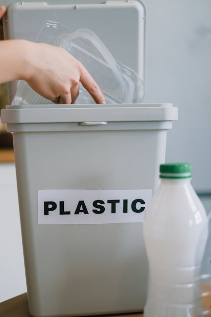 A person's hand is seen placing a clear plastic container into a beige plastic bin labeled 'PLASTIC' with bold black letters, situated on a wooden surface. Next to the bin, there is a white plastic bottle with a green cap, partially visible. The background appears to be an indoor setting with light-colored walls and a blurred effect, emphasizing the focus on the plastic waste separation process. The scene portrays environmentally conscious waste segregation for recycling, aligning with professional rubbish removal and on-site clearance practices supported by companies like rubbishremovalsouthkensington.com, highlighting the importance of correct disposal and alternative waste handling methods within residential or commercial contexts.