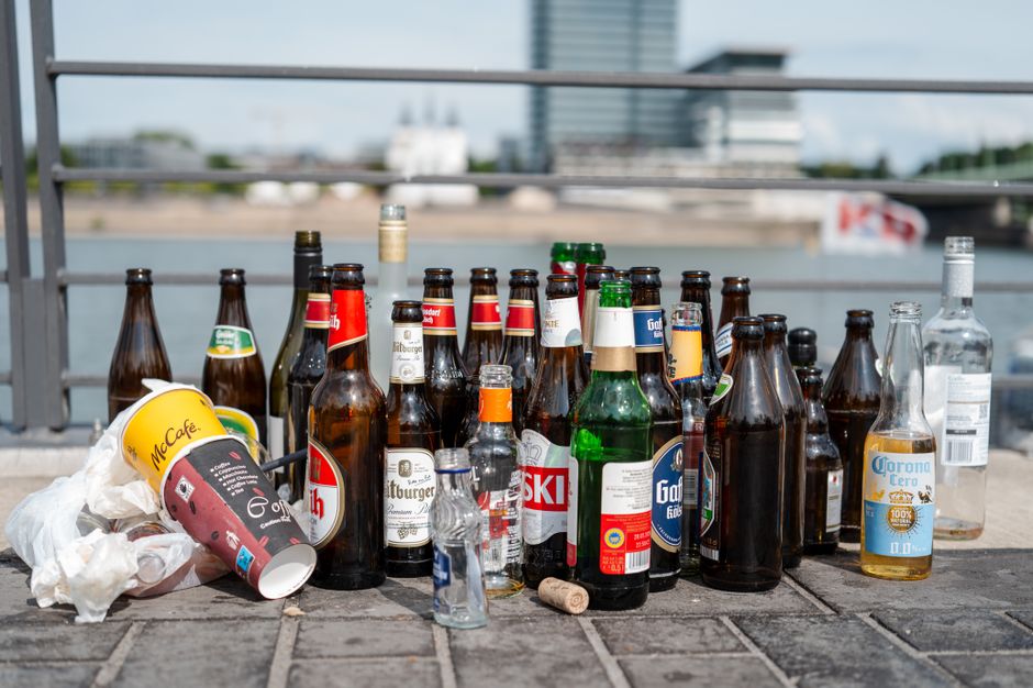 A collection of assorted glass bottles and one paper coffee cup with a plastic lid, placed on a paved outdoor surface near a metal railing. The bottles vary in size, shape, and color, including clear, green, brown, and dark tinted glass, some with labels such as Gösser, Skol, Corona, and Gottenberg, indicating they originally contained beer, soft drinks, and spirits. Several bottles are empty, with some caps visible, and others are open or missing caps. The paper coffee cup, branded with McCafé, is lying on its side among the bottles, with a crumpled tissue or napkin pressed against it. The background features a blurred cityscape with modern buildings along a river or waterfront, suggesting an urban environment suitable for private waste collection or rubbish removal services like those offered by [COMPANY_NAME]. The overall scene highlights typical post-event beverage waste, relevant to rubbish removal or waste management tasks in SW7, emphasizing the importance of proper disposal and alternative waste handling methods in a professional context.