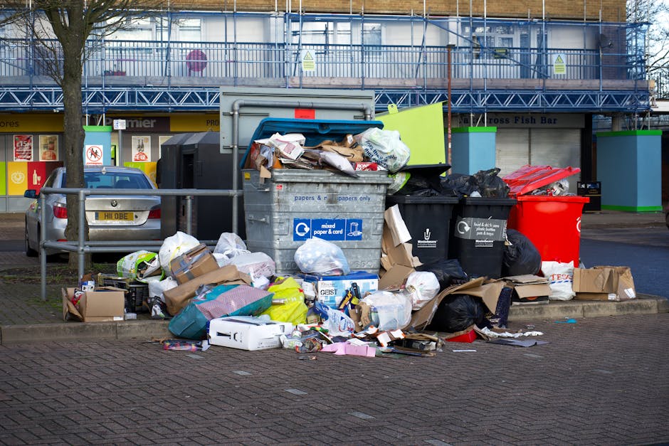 A large assortment of mixed household waste and rubbish is accumulated around a communal recycling bin in an urban outdoor setting, with various types of trash including cardboard boxes, plastic bags, paper, and miscellaneous packaging partially spilling onto the paved pavement and street. The recycling container, made of metal with a blue lid, is designated for paper and cardboard collection, and is surrounded by several smaller black and red waste bins, some of which are overflowing. Behind the waste collection area, a row of storefronts with closed shutters and signage, along with a blue building structure with scaffolding and safety barriers, indicates ongoing construction or maintenance in a busy area. A parked silver car is partially visible to the left, behind a metal railing, and a tall tree trunk is positioned on the left side of the scene. The overall environment suggests a typical city street with waste left for independent rubbish removal services, reflecting the need for on-site clearance or alternative waste handling methods in SW7.