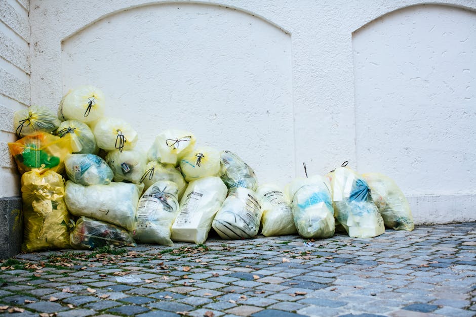 A collection of assorted glass bottles and one paper coffee cup with a plastic lid, placed on a paved outdoor surface near a metal railing. The bottles vary in size, shape, and color, including clear, green, brown, and dark tinted glass, some with labels such as Gösser, Skol, Corona, and Gottenberg, indicating they originally contained beer, soft drinks, and spirits. Several bottles are empty, with some caps visible, and others are open or missing caps. The paper coffee cup, branded with McCafé, is lying on its side among the bottles, with a crumpled tissue or napkin pressed against it. The background features a blurred cityscape with modern buildings along a river or waterfront, suggesting an urban environment suitable for private waste collection or rubbish removal services like those offered by [COMPANY_NAME]. The overall scene highlights typical post-event beverage waste, relevant to rubbish removal or waste management tasks in SW7, emphasizing the importance of proper disposal and alternative waste handling methods in a professional context.
