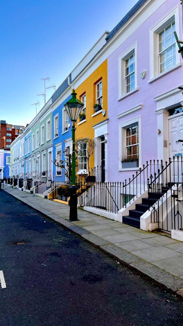 A row of colorful terraced houses in pastel shades of blue, yellow, and lavender along a narrow urban street. Each property features small front steps leading up to white-framed windows and doors, with some houses having flower boxes beneath the windows. In the foreground, a black metal street lamp with a classic design is positioned on the sidewalk, which is paved with grey slabs. To the left, there is a black wrought iron fence enclosing small front gardens or steps. The street surface is dark asphalt, and the sky is clear, suggesting a bright day. The scene reflects a typical residential area with well-maintained facades, illustrating an environment where private waste management or rubbish removal services might operate for property clearance or routine collection, set against a backdrop of vibrant neighbourhood aesthetics.