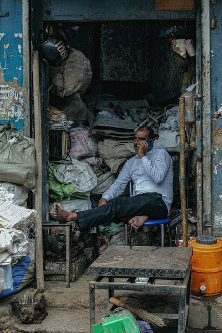 A man seated on a small, black, patterned stool inside a cluttered storage space or container, surrounded by large bags of mixed waste materials such as plastic, paper, and textiles. He is wearing a light blue shirt and dark pants, holding a mobile phone to his ear, with a neutral facial expression. The environment appears cramped with an assortment of loose items, tools, and containers, including an orange vacuum or blower on the right side and a small, green object on the ground in front of the stool. The back wall of the space is dark, with some shelves or stacked materials, suggesting a setting related to waste collection or disposal. The scene is lit with natural or ambient light, emphasizing the functional, utilitarian nature of the area, which may be associated with independent rubbish handling or private waste management activities, illustrative of alternative rubbish removal scenarios sometimes encountered in urban environments.