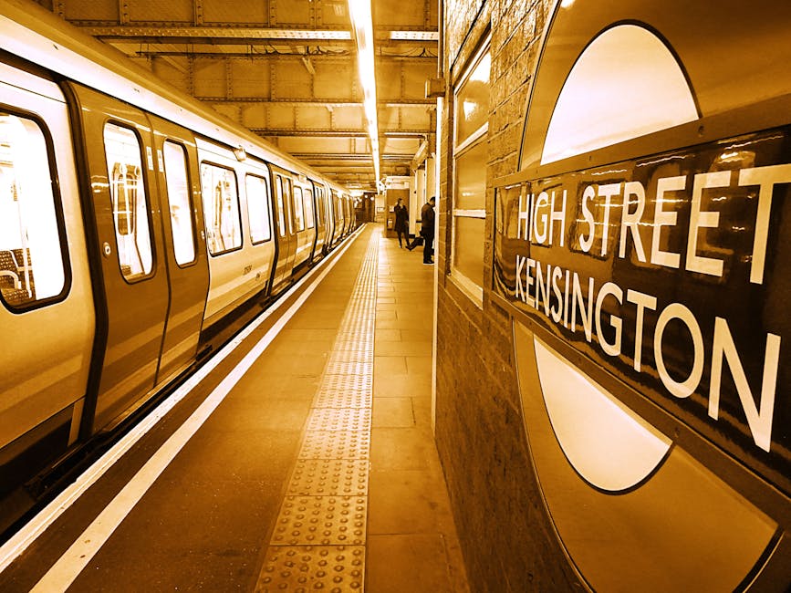 A photograph of an underground London Underground station platform at High Street Kensington, showing a London Underground train with a cream and dark blue exterior parked alongside. The train features rectangular windows, some with visible reflections, and closed doors. The platform is illuminated by overhead strip lighting, casting a warm tone across the scene. The surface of the platform is tiled with a central tactile paving strip for visually impaired passengers. To the right, part of a brick wall with a rounded edge is visible, featuring a large, round London Underground roundel sign with a white background, black border, and white text reading 'HIGH STREET KENSINGTON.' A few passengers are standing further down the platform, some facing the train. The environment appears clean and well-maintained, with no visible rubbish, consistent with a professional waste management context represented by Rubbish Removals South Kensington and sustainable waste handling services.