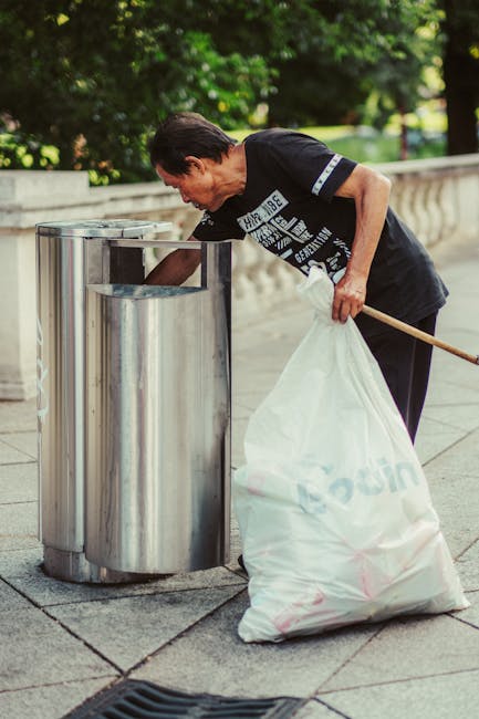A narrow urban alleyway shows a large grey and black rolling rubbish cart positioned in the center foreground, partially covered with a weathered fabric cover. Behind the cart, stacked piles of cardboard boxes, paper bags, and other waste materials are seen against a backdrop of general debris and discarded items. The alley is flanked by tall buildings on both sides, with graffiti visible on one side and a mixture of brick and painted surfaces on the other. Bare tree branches extend overhead, indicating a cold or late autumn season. The ground is uneven and stained with patches of dirt and grime, suggesting it is used for waste disposal or debris collection. Ambient natural lighting emphasizes the textured surfaces and litter, reflecting a typical scene where independent rubbish collection services, such as those provided by [COMPANY_NAME], might perform on-site clearance for local residents or businesses. The overall atmosphere conveys a utilitarian area designated for waste storage and disposal within an urban environment.