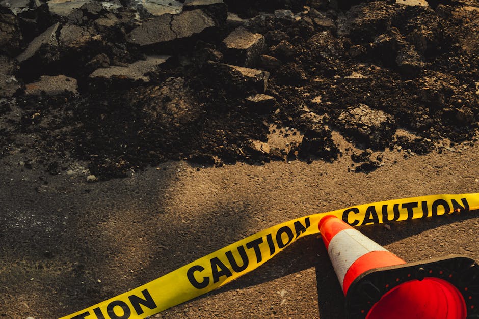 Close-up view of a construction or maintenance site showing a section of asphalt pavement with uneven, broken asphalt pieces and loose gravel. The foreground features a yellow caution tape with black text reading 'CAUTION' stretched across the scene, attached to an orange and white traffic cone lying on its side. There is also a bright orange warning light resting on the ground near the cone, indicating an ongoing or restricted work area. The environment appears outdoor with natural lighting, and the disrupted pavement suggests recent excavation or repair work likely related to underground utility or road maintenance, aligning with the context of waste management and site clearance services offered by companies like rubbishremovalsouthkensington.com.