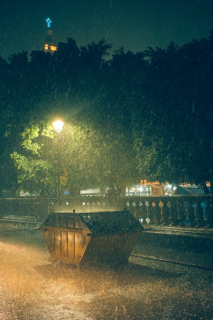 The image depicts a street scene during a heavy rainstorm at night, with water splashing on the pavement and overflowing from a large, brown, wooden skip positioned on the roadside, partially filled and tilted slightly forward. Behind the skip, there is a metal railing separating the street from a sidewalk with leafy trees, whose branches cast shadows and diffuse the street lighting. A traditional street lamp emits a warm glow, illuminating the wet surface and creating reflections on the damp pavement. In the background, there is a building with exterior lighting and a tall structure topped with a lit cross, visible through the rain and trees. The overall scene captures the weather conditions impacting outdoor waste collection or disposal activities, illustrating how private rubbish removal services such as those from rubbishremovalsouthkensington.com might operate in adverse weather, with a focus on safety and practicality during on-site clearance or rubbish collection.