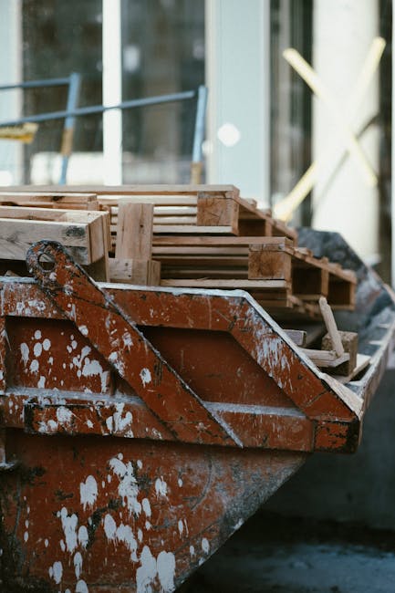 A close-up view of a rusted, orange-brown metal boat or trolley used for construction or waste collection, fitted with chipped white paint and streaks of white residue. Resting on a concrete or paved surface, the boat is loaded with an assortment of wooden pallets and planks, each varying in size and color, with some showing signs of weathering and rough texture. In the background, the scene features a building with large glass windows reflecting natural daylight, alongside blurred outdoor elements such as a wooden garden feature or fence. The overall environment suggests an outdoor workspace or storage area associated with construction or waste management activities, potentially related to independent rubbish removal services. The presence of wooden waste alongside metal equipment emphasizes the need for appropriate disposal or recycling of mixed materials, aligning with the themes of private waste handling and alternative rubbish collection methods, as managed by rubbishremovalsouthkensington.com.