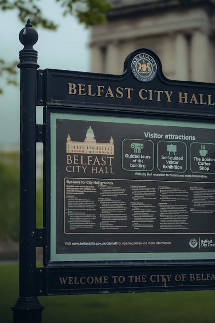A large, dark blue information sign positioned outdoors in front of Belfast City Hall, featuring the official Belfast City Council seal at the top. The sign displays the header 'BELFAST CITY HALL' in gold lettering and incorporates multiple sections of text and icons. The main contents include details about visitor attractions, such as guided tours, self-guided exhibitions, and the Bobbin Coffee Shop. The lower part contains a block of detailed guidelines and bye-laws regarding city hall grounds, with small, densely packed text in white on a dark background. The environment around the sign includes a blurred view of greenery and a portion of the historic city hall structure with classical architecture, visible in the background. The lighting appears natural, with soft daylight illuminating the scene, and the sign is mounted on a decorative post, emphasizing its role as an informational fixture for visitors.